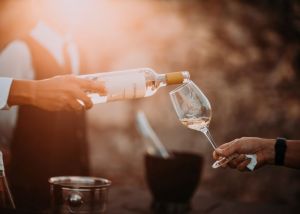 Pessoa enche a taça de vinho da amiga com vinho branco em local aberto durante pôr do sol